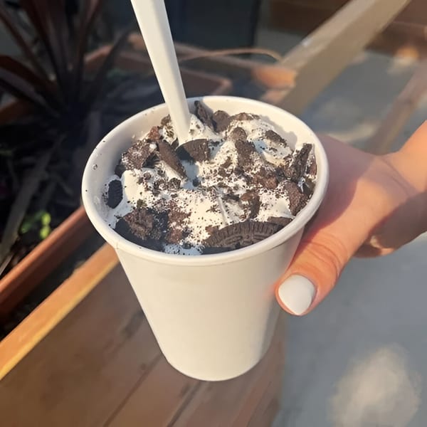 A hand holds a white cup filled with creamy dessert topped with crushed chocolate cookies and a spoon.