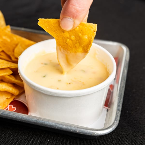 Hand dipping a tortilla chip into a cup of creamy BTB Burrito queso, with more chips nearby.