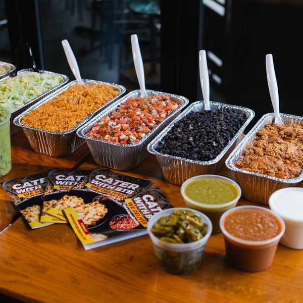 Catering spread with aluminum trays of rice, beans, salsa, and meat, alongside sauces and catering flyers.