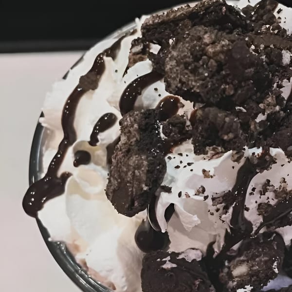 Close-up of a dessert with whipped cream, chocolate syrup, and crumbled cookies in a glass.