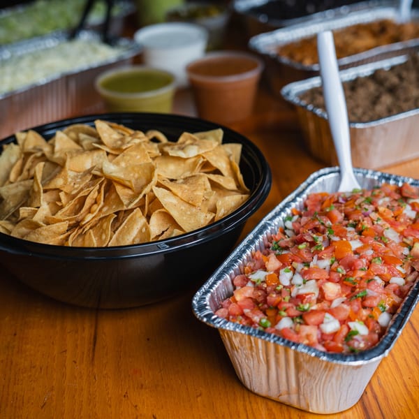 A black bowl of tortilla chips and a foil tray of pico de gallo on a wooden table.