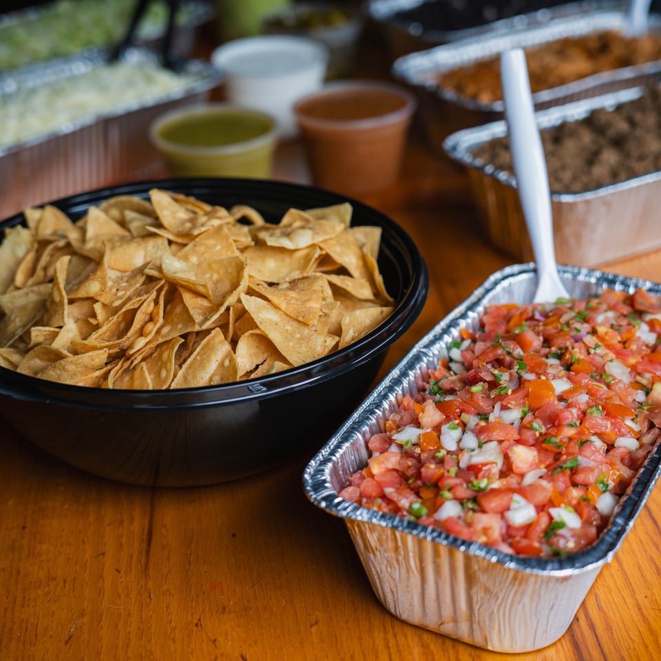 A black bowl of tortilla chips and a foil tray of pico de gallo on a wooden table.