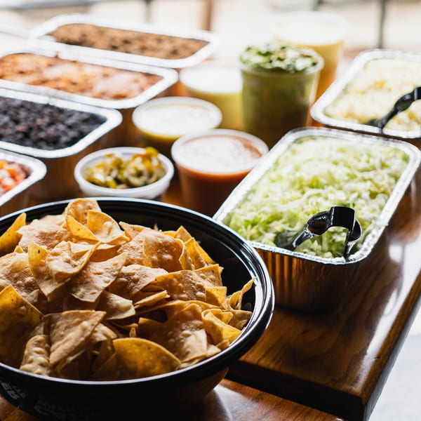 A buffet spread of Mexican food, including chips, beans, rice, and various toppings in foil containers.